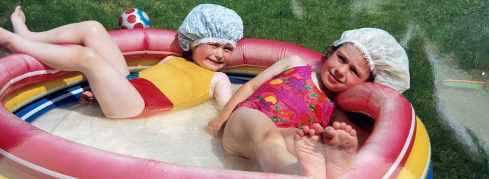 two young girls wearing showercaps and lounging in a paddle pool on the grass
